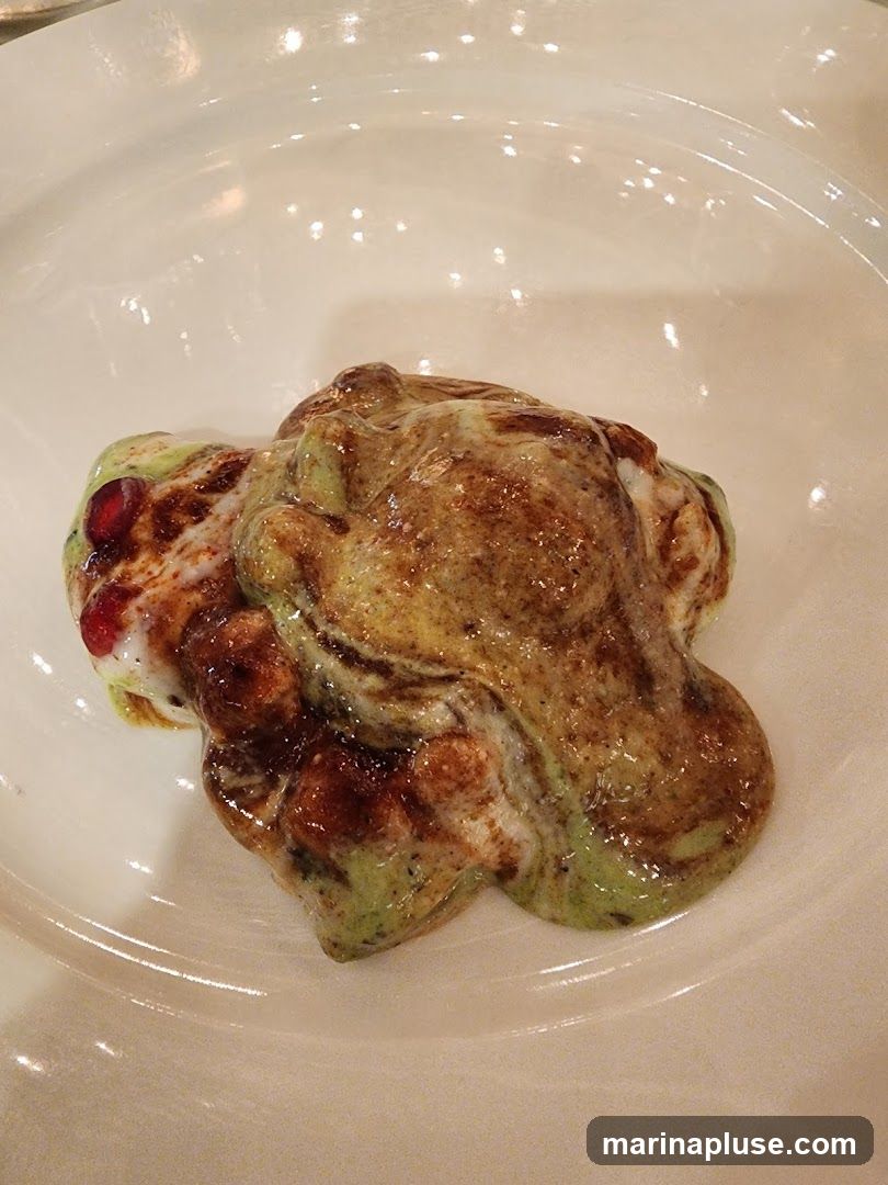 A close-up shot of a savory Indian snack served on a white plate. The dish appears to be a chaat, with layers of yogurt, chutneys, and crispy elements. Garnished with pomegranate seeds and spices, it has a vibrant and textured appearance.
