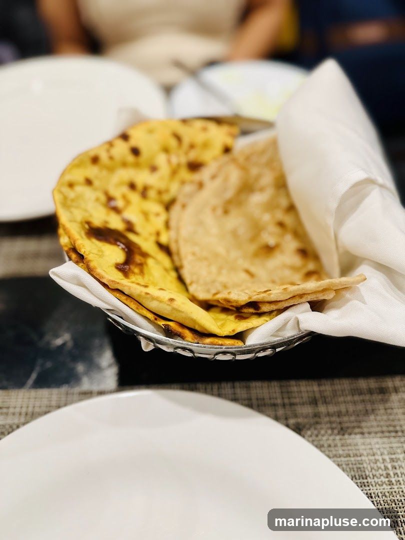 A close-up shot of a metal basket filled with Indian breads, including what appears to be naan and roti. The breads are partially covered by a white cloth napkin. The basket sits on a dark, textured surface, with white plates visible in the foreground and blurred figures in the background, suggesting a dining setting.