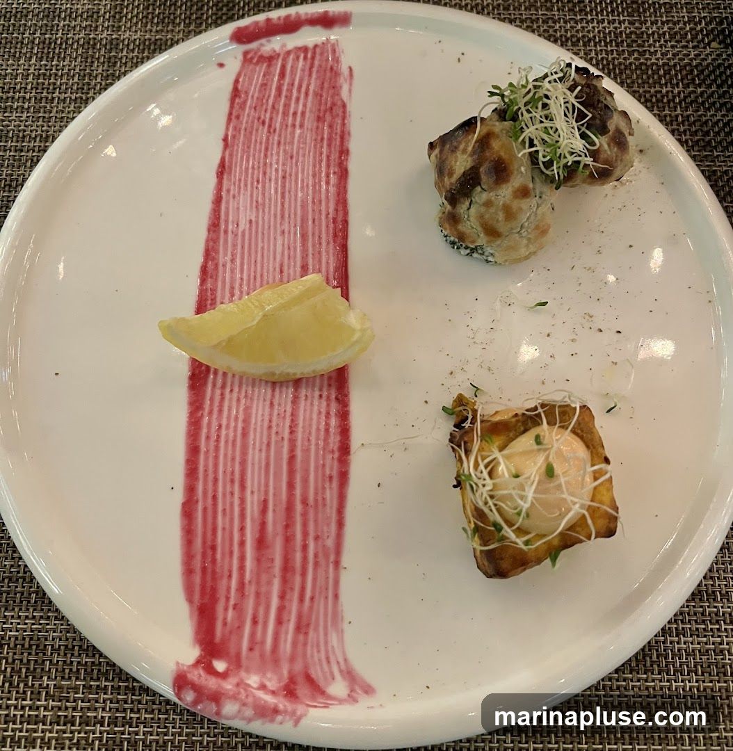 A white plate with a red sauce smear, a lemon wedge, and two small appetizers. One appetizer appears to be a pastry filled with a creamy substance and sprouts. The other appetizer consists of two small, round items topped with sprouts.