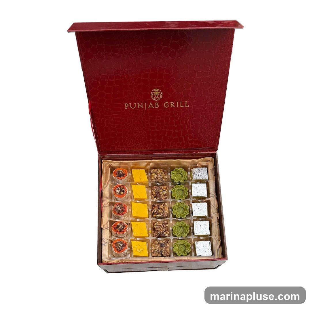 A red, textured gift box with the logo and name "PUNJAB GRILL" embossed in gold on the lid. The box is open, revealing an assortment of Indian sweets arranged in individual compartments. The sweets include circular, orange-colored items with nuts, yellow square pieces, square pieces with nuts and dried fruit, green flower-shaped sweets, and silver-wrapped square pieces.