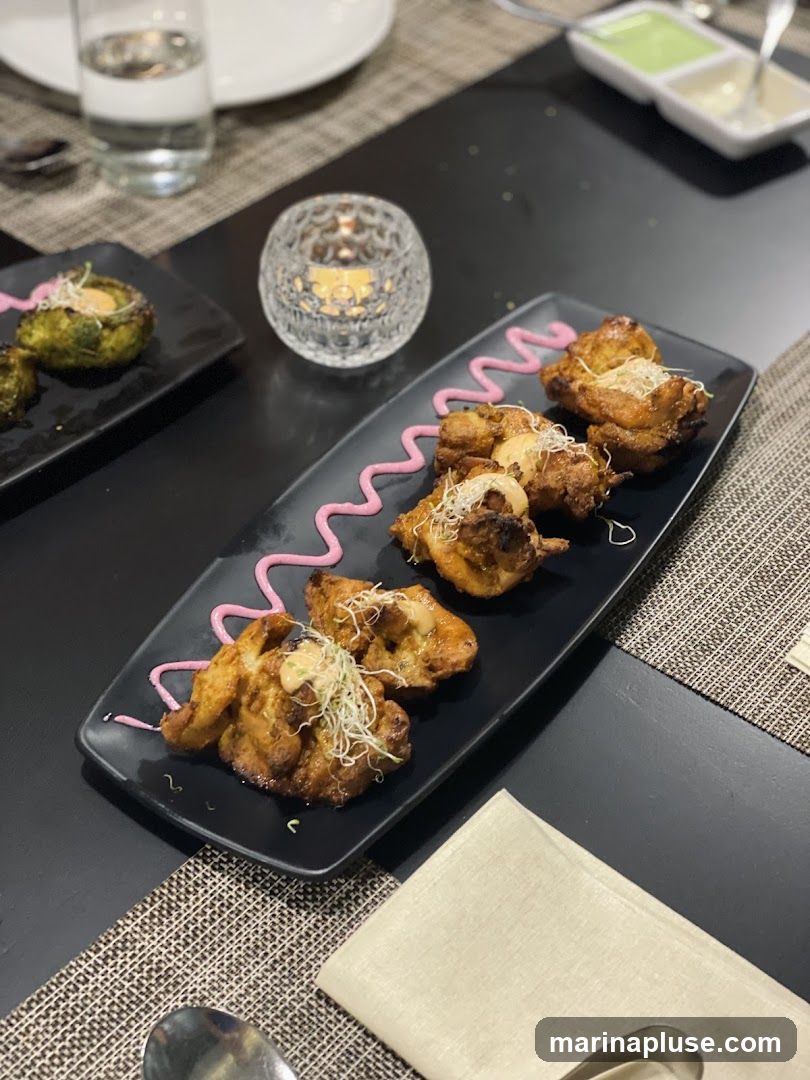 A close-up shot of two black plates with appetizers, a glass of water, and a candle in a decorative holder on a table. The plate in the foreground contains several pieces of grilled chicken or paneer, garnished with sprouts and a pink sauce. The plate in the background has green appetizers, also garnished. Two small white dishes with dips are visible in the upper right corner.