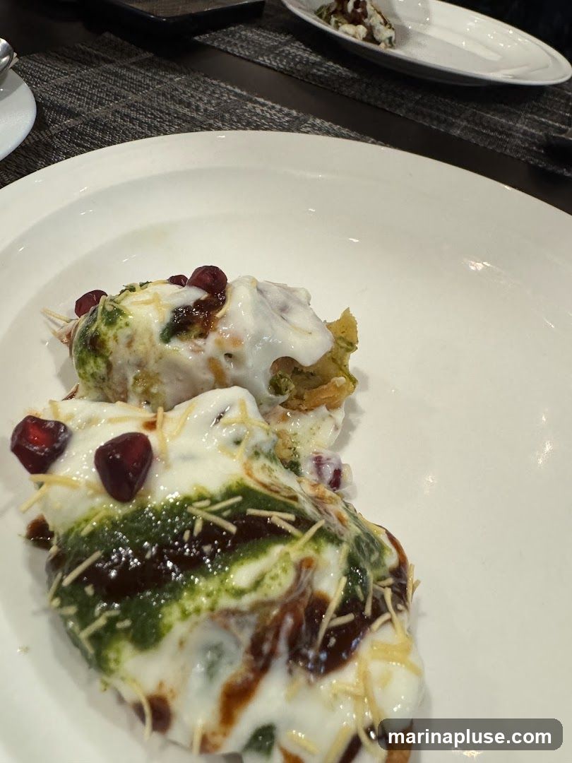 A close-up shot of a plate of Indian street food, likely a type of chaat. The dish is topped with yogurt, green chutney, tamarind sauce, pomegranate seeds, and sev (crispy chickpea noodles). The food is presented on a white plate, with another plate of the same dish visible in the blurred background. The setting appears to be a restaurant with a dark, textured placemat on the table.