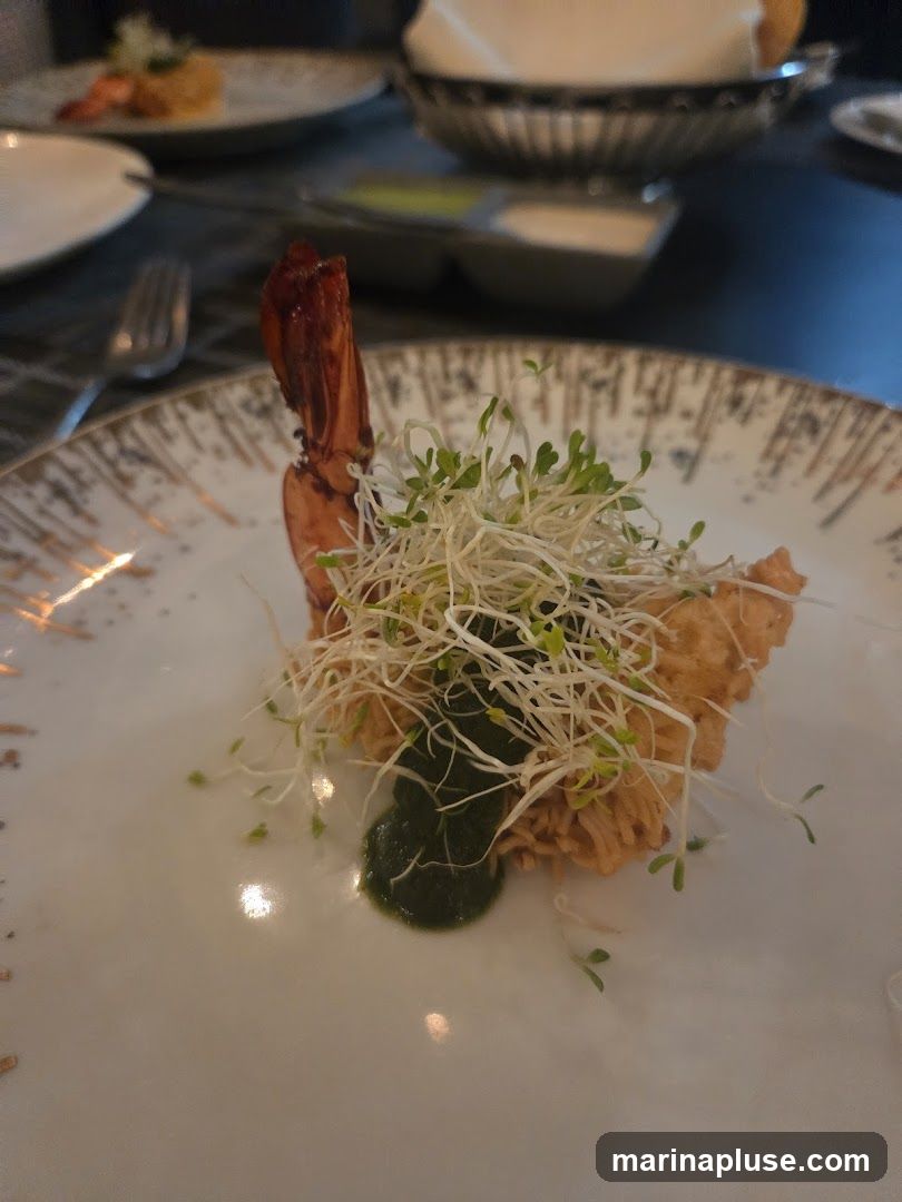 A close-up shot of a single fried shrimp appetizer on a white plate. The shrimp is topped with a generous portion of sprouts and a dollop of green sauce. The plate has a decorative gold rim. In the background, other plates with food and a basket of bread are visible, suggesting a restaurant setting.
