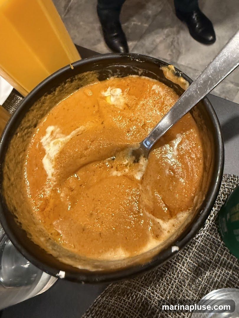 A close-up overhead view of a bowl of creamy orange curry, likely a Indian dish, with a swirl of white cream on top. A silver spoon is resting in the bowl. To the left, a glass of orange juice is visible. In the background, the legs of two people wearing dark shoes and pants are visible. The setting appears to be a restaurant.