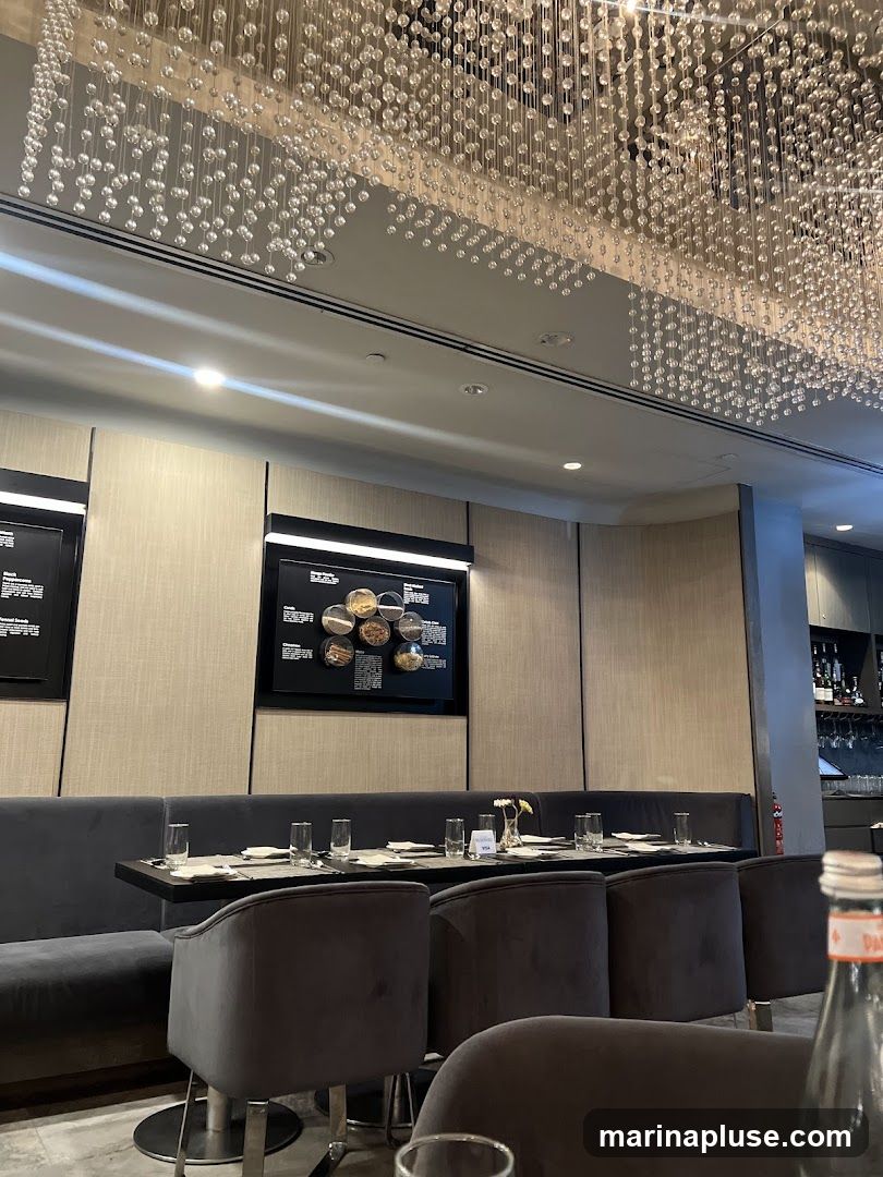 The image shows the interior of a restaurant with a long, plush banquette seating area. The tables are set with glassware and napkins. Above, a striking chandelier made of numerous clear spheres hangs from the ceiling, reflecting light. On the wall, there are framed displays, one of which appears to show various spices or ingredients in small bowls. The overall ambiance is sophisticated and modern.