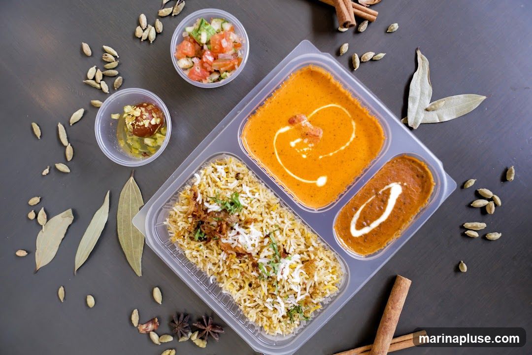 A top-down view of a takeaway container filled with Indian food. The container has three compartments: one with biryani, another with a creamy orange curry, and a third with a dark lentil dish. There are also two small side containers, one with a salad and the other with a dessert. The food is arranged on a dark surface, with scattered spices like cardamom pods, bay leaves, and cinnamon sticks around it.