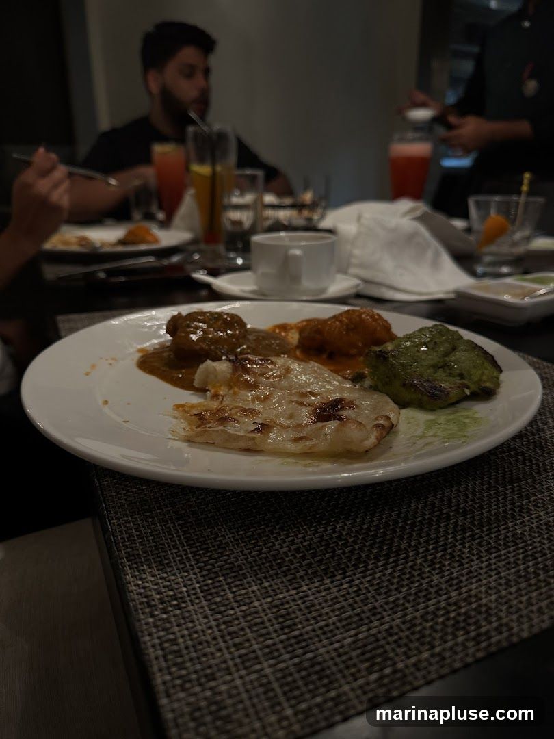 A close-up shot of a white plate filled with various Indian dishes, including what appears to be tandoori chicken, a curry, and naan bread. The food is arranged on a dark, textured tablecloth. In the blurred background, diners are seated at tables with drinks, suggesting a restaurant setting.