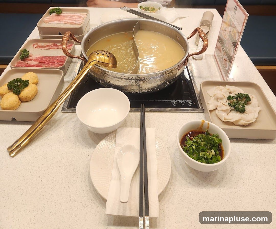 A table set for a hot pot meal with a split pot of broth, various raw ingredients like thinly sliced meats and fish balls, dumplings, a dipping sauce, and dining utensils. A menu stand is visible in the background.