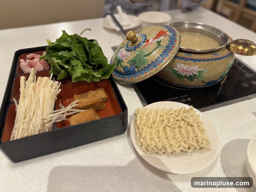 A close-up view of a hot pot meal setup. In the foreground, a black tray contains thinly sliced meat, leafy green vegetables, enoki mushrooms, and fried tofu. To the right, a small white plate holds a block of instant noodles. A decorative hot pot with a floral design sits on a heating element, filled with broth.