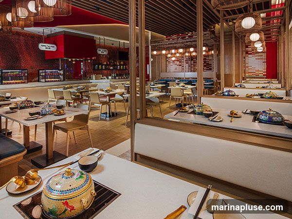 An interior view of a modern restaurant with tables set for dining. The tables are equipped with individual cooking stations, suggesting a hot pot or similar dining experience. The decor features a mix of wood, red accents, and ambient lighting, creating a warm and inviting atmosphere. In the background, there are shelves stocked with bottles and what appear to be menu boards or displays.