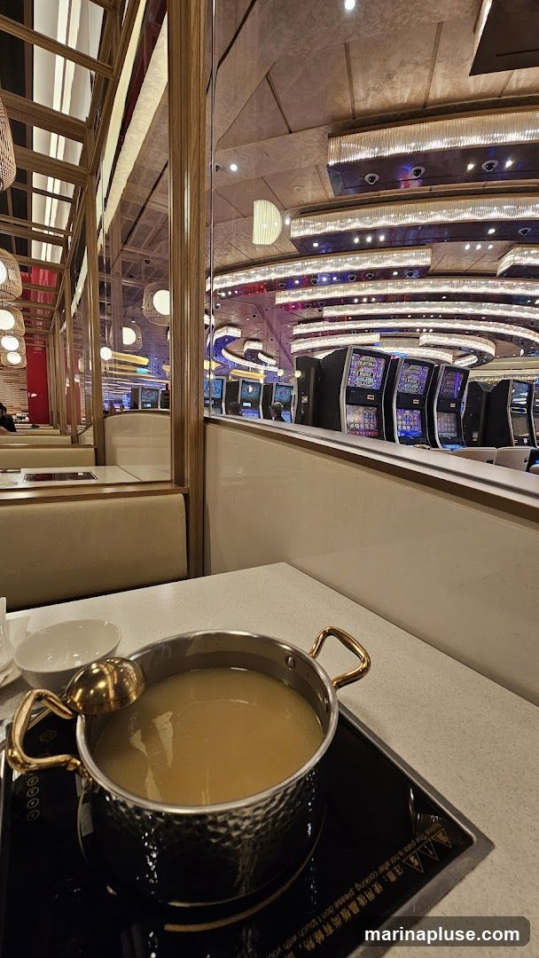 A close-up view of a pot of broth on an induction cooktop, with a golden ladle resting on the rim. In the background, through a reflective partition, a row of slot machines in a casino is visible, illuminated by colorful lights. The dining area features booth seating with wooden accents and decorative lighting.
