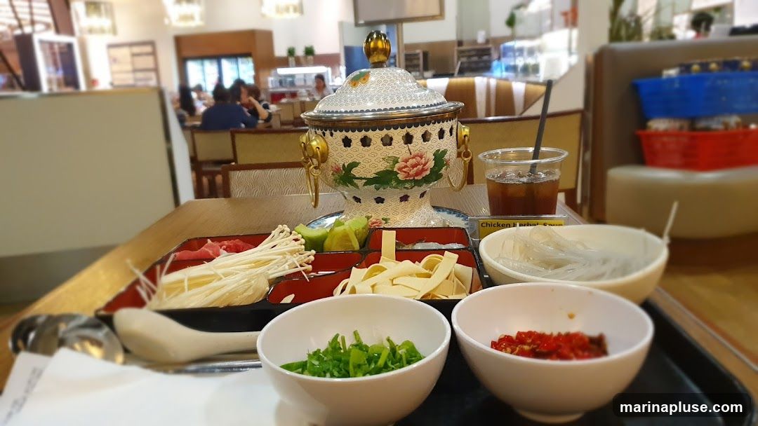 A table setting for hot pot with a decorative pot, a tray of thinly sliced meat and enoki mushrooms, sliced tofu, and a bowl of chopped herbs and a bowl of chopped chilies. A drink with ice and a straw is also visible, along with a label that reads "Chicken Herbal Soup". In the background, other diners are seated at tables in a restaurant setting.