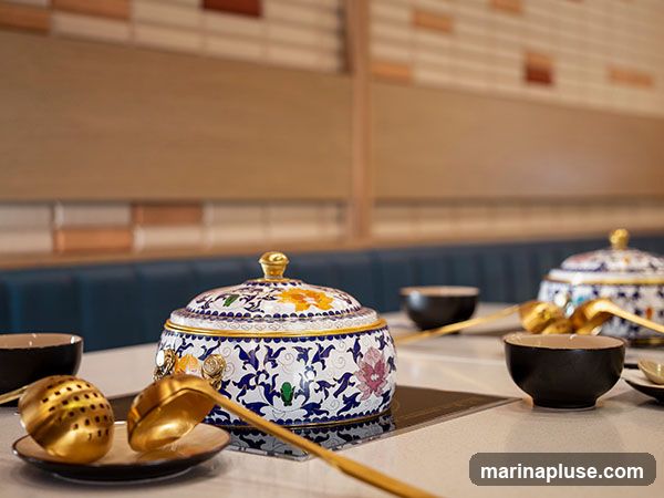 A close-up view of a table set for a hot pot meal. In the foreground, a decorative lidded pot with intricate blue and white floral patterns and yellow accents is placed on a heating element. Beside it are golden ladles and a slotted spoon. Several dark bowls are arranged on the table. In the background, another similar lidded pot and bowls are visible, suggesting a dining setting. The wall behind the table features a tiled pattern of light brown and reddish-brown rectangles.