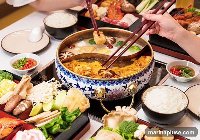 A close-up shot of a hot pot meal with various ingredients. In the center, a split pot contains two different broths, one clear and one reddish-orange. Chopsticks are seen dipping into the pot, picking up pieces of food. Around the pot, there are platters filled with fresh ingredients such as corn on the cob, enoki mushrooms, broccoli, shiitake mushrooms, leafy greens, and thinly sliced meat. Bowls of white rice and dipping sauce are also visible on the table. The scene suggests a communal dining experience.