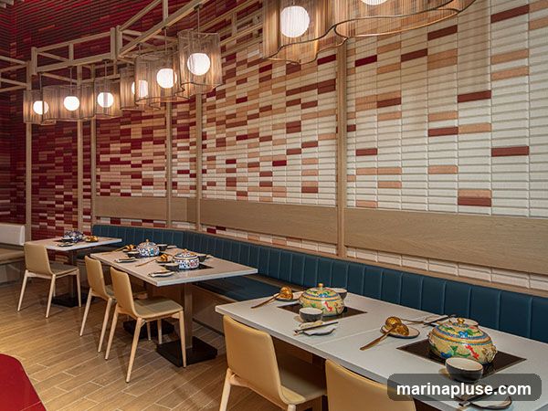 The interior of a restaurant features tables set up for hot pot dining. The walls are decorated with a mosaic of rectangular tiles in shades of red, cream, and beige. Modern pendant lights with cylindrical shades hang from a wooden frame above the tables. The tables are white, with built-in induction cooktops. Blue upholstered banquettes line the walls, with individual chairs at the tables. Each place setting includes bowls, chopsticks, and a small dish. Decorative hot pot vessels are placed on the cooktops.