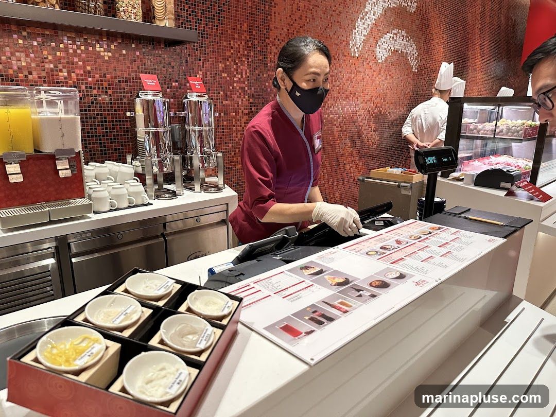 A food service counter with a staff member wearing a mask and gloves serving a customer. There are dispensers for drinks, cups, and a display of small bowls with various toppings. A menu is laid out on the counter, showing pictures of food items. In the background, another staff member in a chef's hat is visible near a display case with food. The wall behind the counter is decorated with a mosaic tile pattern.