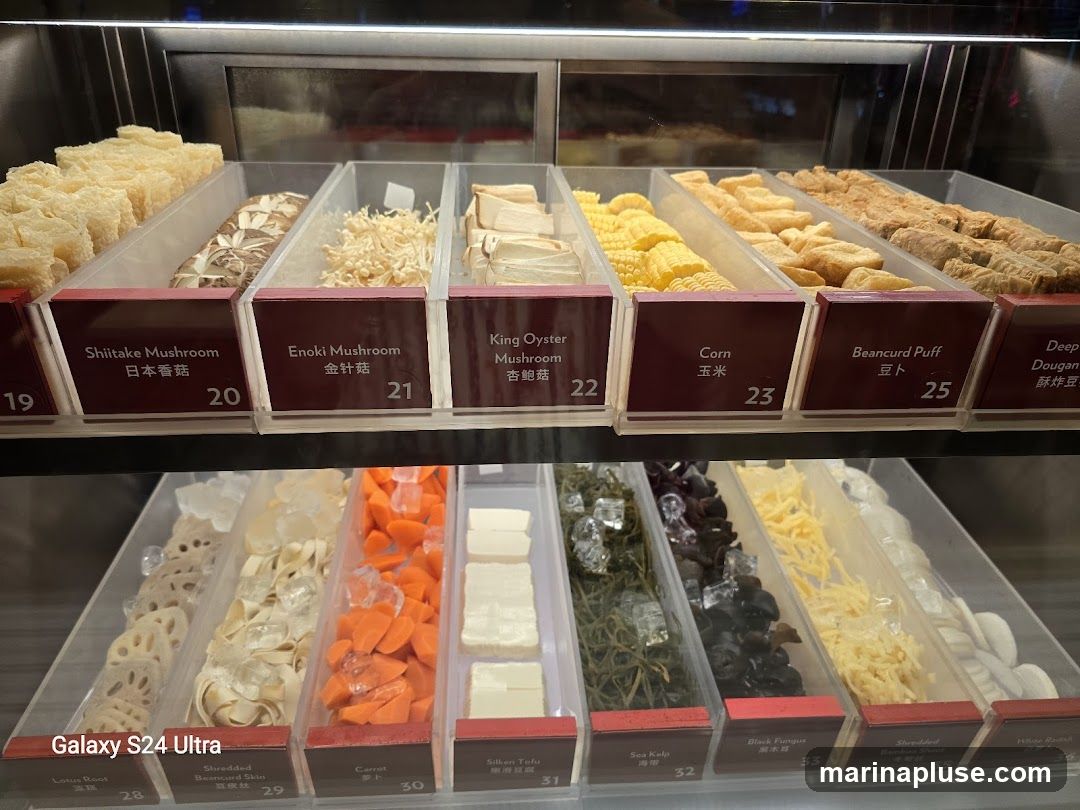 A display case filled with various ingredients, likely for hot pot or a similar dish. The ingredients are neatly arranged in clear containers with labels. Visible items include different types of mushrooms (Shiitake, Enoki, King Oyster), corn, beancurd products, lotus root, shredded beancurd skin, carrots, sea kelp, black fungus, shredded bamboo shoots, and white radish.