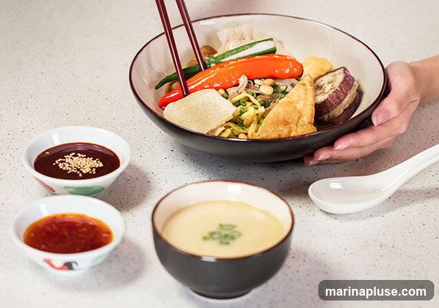 A close-up shot of a bowl of noodles with various toppings, including red peppers, cucumber, eggplant, and fried tofu. Two small dipping bowls, one with a dark sauce and sesame seeds, and another with a red chili sauce, are placed to the left of the main bowl. A bowl of creamy soup with green onions and a white spoon are on the right. Chopsticks are being used to pick up food from the noodle bowl.
