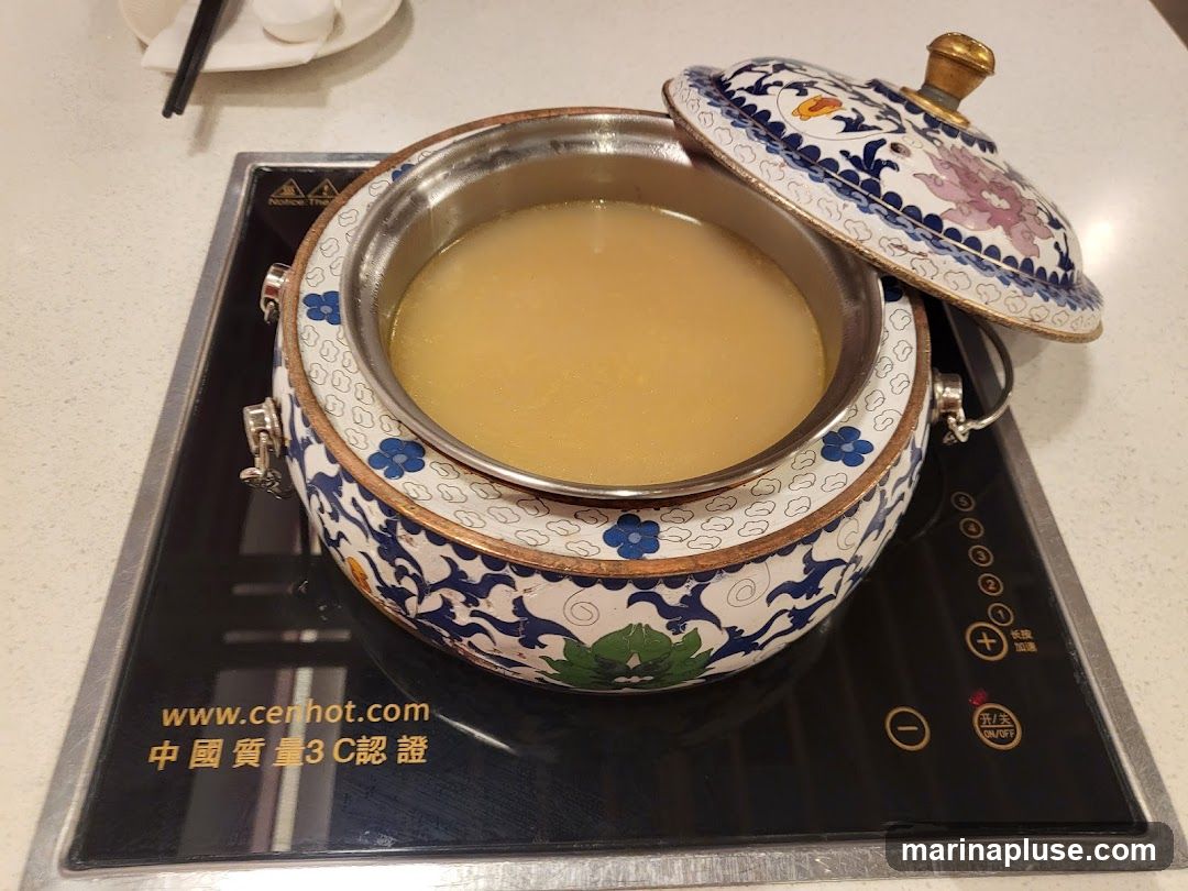 A decorative pot filled with light-colored broth sits on an induction cooktop. The pot has intricate blue and white floral patterns and a matching lid. The cooktop has visible controls and a website address printed on it.