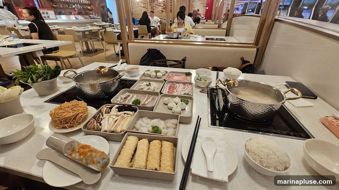 A table set for hot pot dining with various ingredients laid out. In the foreground, there are bowls of rice, a plate of fried items, and a dish with what appears to be fish roe and paste. Behind these, several trays hold thinly sliced meats, meatballs, mushrooms, and noodles. Two pots are on induction burners, one with simmering broth. In the background, other diners are seated at tables in a restaurant setting.
