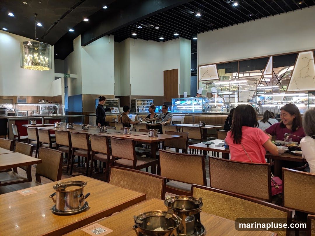 A view of a restaurant interior with tables and chairs set up for dining. Several patrons are seated at tables, and staff members are visible in the background. The restaurant appears to have a buffet or display counter with food items.