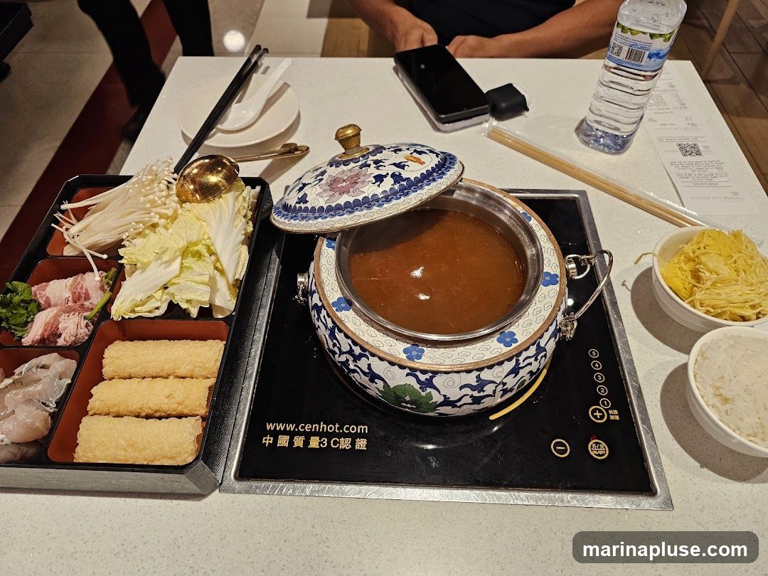 A hot pot meal is laid out on a table, featuring a decorative pot filled with broth, a tray of raw ingredients including thinly sliced meat, fish, enoki mushrooms, and napa cabbage, along with fried tofu sticks. Two bowls, one with shredded egg and another with rice, are also visible. A bottle of water and receipts are on the table. The induction cooker has the website www.cenhot.com printed on it.