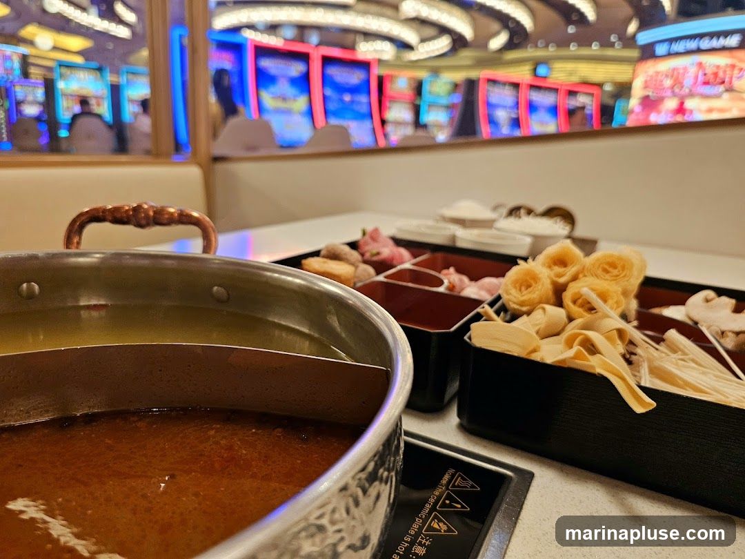 A close-up view of a hot pot with two broths, one clear and one reddish-brown, sits on a table. In front of the pot, a black tray holds various ingredients for the hot pot, including thinly sliced meat, tofu skin, enoki mushrooms, and what appear to be flower-shaped tofu or fish cakes. In the background, blurred slot machines with bright lights and screens are visible, suggesting the dining area is within a casino.