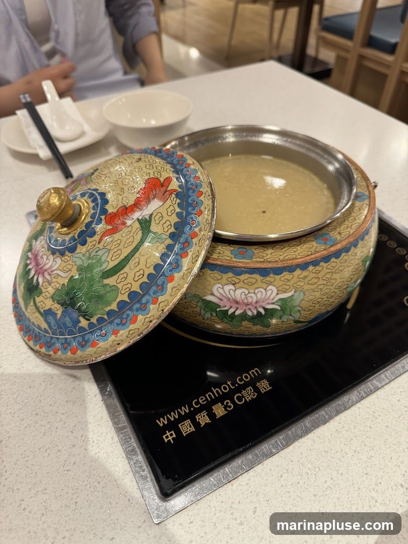 A decorative hot pot dish with a lid, filled with a light-colored broth, sits on a black induction cooktop. The dish is adorned with floral patterns in blue, red, and green. A white bowl and chopsticks are visible on the table in the background. The cooktop has text that includes a website address and Chinese characters.