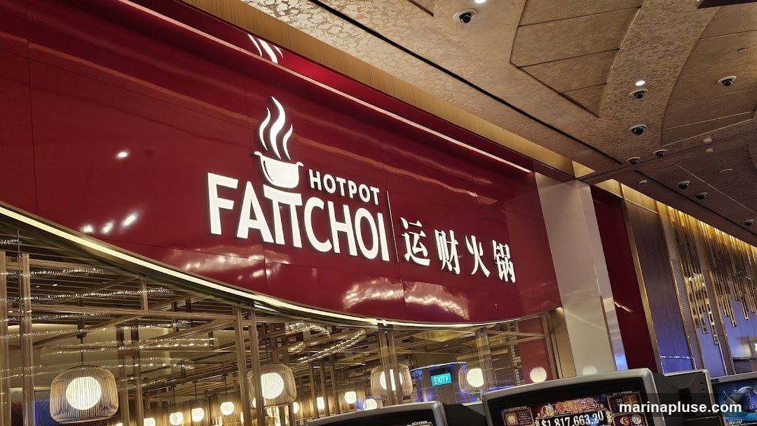 A close-up shot of a red sign with white lettering that reads "HOTPOT FATTOI" in English and Chinese characters. The sign is illuminated and appears to be above the entrance to a restaurant. Below the sign, there is a glimpse of the interior with decorative lighting and what appear to be slot machines in the foreground.