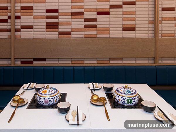 A white table is set for a hot pot meal with two decorative ceramic pots, bowls, chopsticks, and ladles. The background features a wall with a textured pattern of rectangular tiles in shades of beige, brown, and red, with a wooden accent panel and blue upholstered seating.
