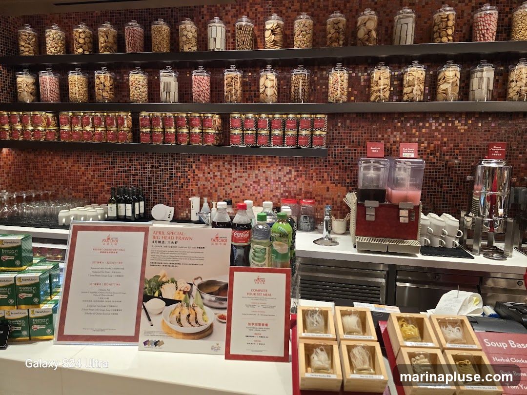 A restaurant counter display featuring various food items and promotional materials. Shelves above are stocked with jars of snacks and cans of food. The counter displays drinks, a soup base selection, and various types of noodles. Two promotional signs are prominently displayed, one detailing an April special for "Big Head Prawn" and another for a "Complete Set Meal."