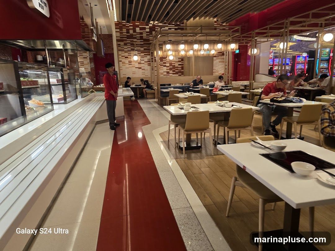 A view of a restaurant dining area with tables set for hot pot. A staff member in a red shirt stands near a display counter. Diners are seated at various tables, some eating. The decor features wooden accents and red and white color schemes. In the background, slot machines are visible.
