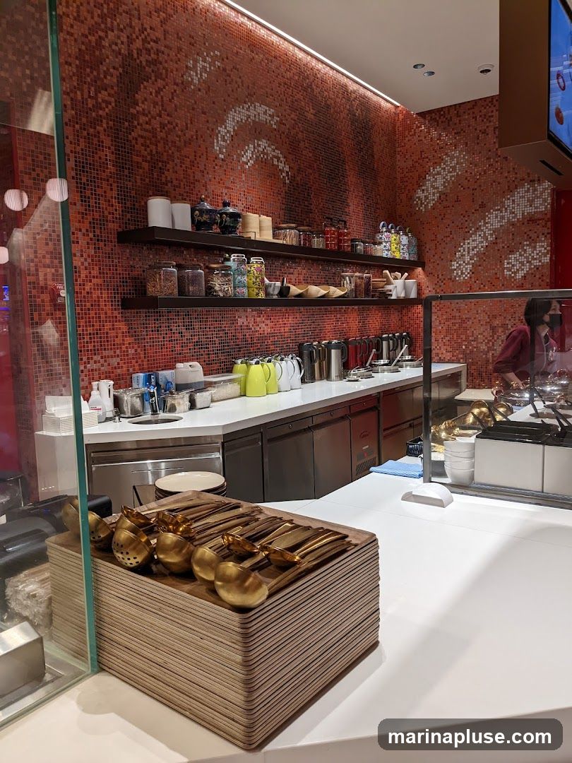 A close-up view of a clean and modern kitchen counter area, featuring stacks of wooden trays topped with golden serving spoons and ladles. Behind the counter, shelves are stocked with various jars and containers, and a tiled wall with a decorative pattern adds a vibrant touch. A person in the background is visible near a food service station.