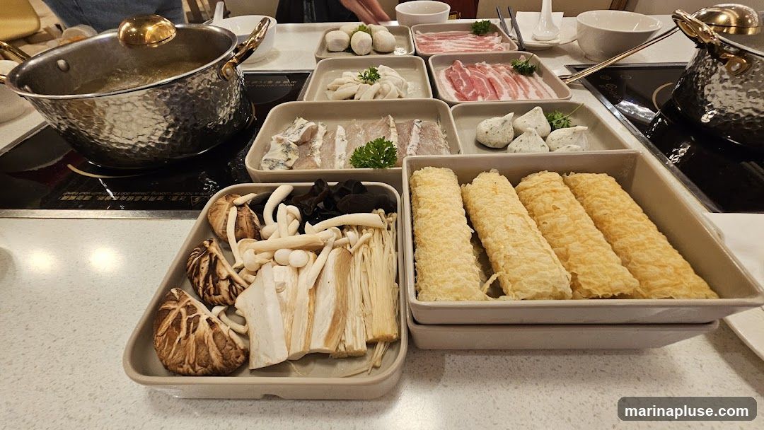 A table set for hot pot with various ingredients laid out in rectangular dishes. In the foreground, a dish contains an assortment of mushrooms, including shiitake and enoki, along with black fungus. To the right, a dish holds several fried tofu skin rolls. Behind these, other dishes contain thinly sliced meat, fish slices, and various types of meatballs. A pot of broth simmers on an induction cooker on the left side of the image.