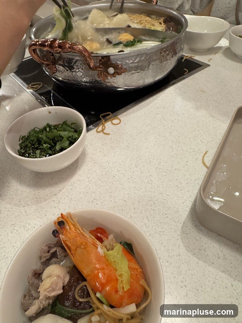 A close-up view of a hot pot meal with various ingredients. In the foreground, a bowl contains a large shrimp, thinly sliced meat, and noodles. Behind it, a communal hot pot is simmering with broth, vegetables, and noodles. A small bowl of chopped herbs is also visible on the table.
