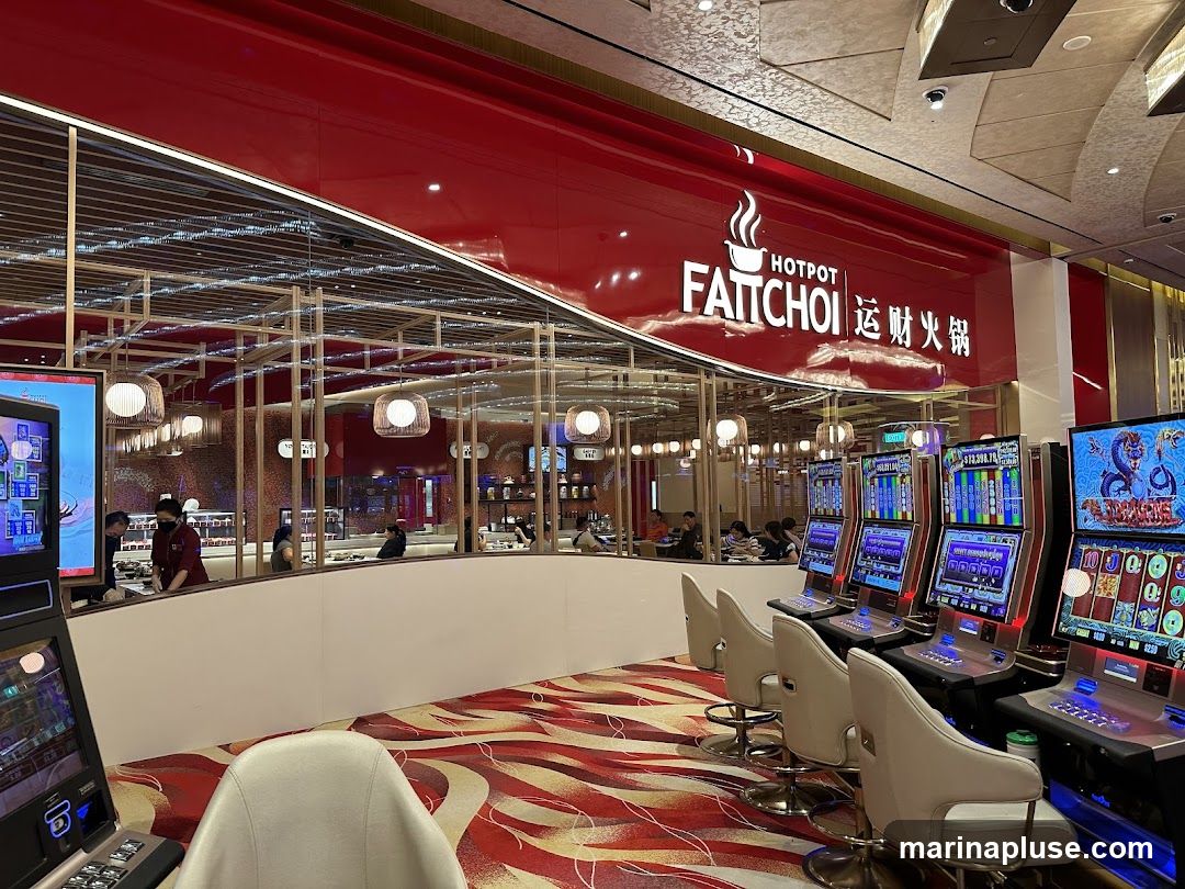 A brightly lit interior with a red and gold color scheme. In the foreground, several slot machines are lined up, with chairs in front of them. Behind the slot machines, a hot pot restaurant is visible through a glass partition. The restaurant has a modern design with wooden accents and hanging lamps. The sign for the restaurant, "HOTPOT FATCHOI," is prominently displayed in white lettering against a red background. People are seated at tables inside the restaurant. The floor is covered with a red and white patterned carpet.
