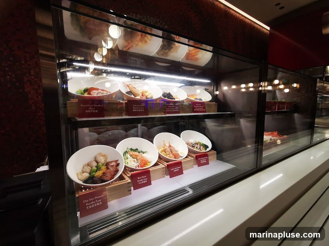 A display case filled with various food items, including roast duck, noodles, seafood, and dumplings. Each dish is presented in a white bowl and labeled with its name in English and Chinese. The display is well-lit, highlighting the appetizing presentation of the food.