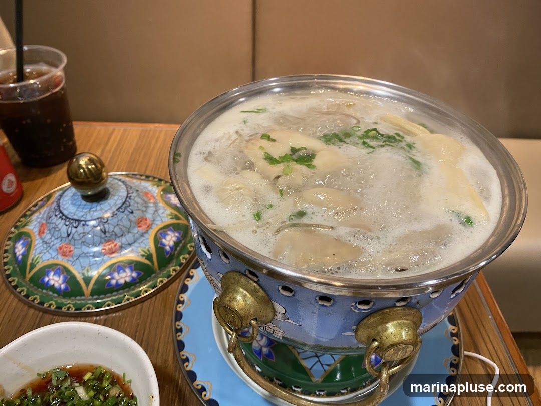 A close-up shot of a hot pot dish with dumplings and broth, garnished with green onions. A decorative lid and a small bowl of dipping sauce are visible on the side, along with a cup of dark beverage with a straw. The meal is served on a wooden table.