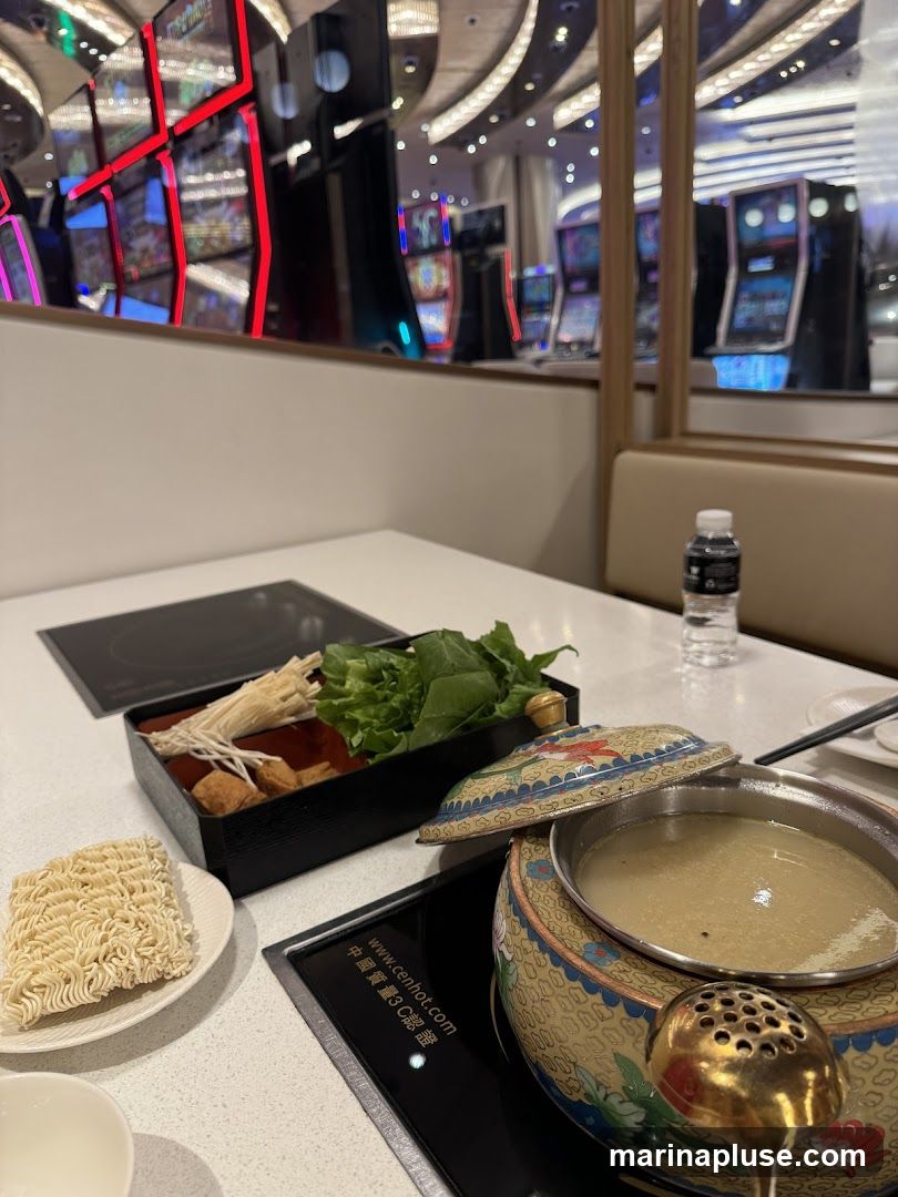 A table set for hot pot with a pot of broth, a plate of instant noodles, and a tray of vegetables and other ingredients. In the background, rows of brightly lit slot machines are visible, suggesting the dining area is within a casino or entertainment venue. A bottle of water is on the table.