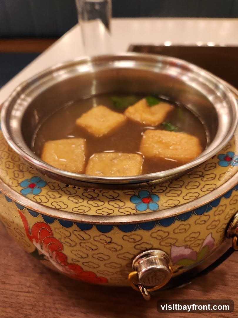 A close-up shot of a small metal pot filled with broth and several pieces of fried tofu. The pot is decorated with a yellow base and blue and red floral patterns.