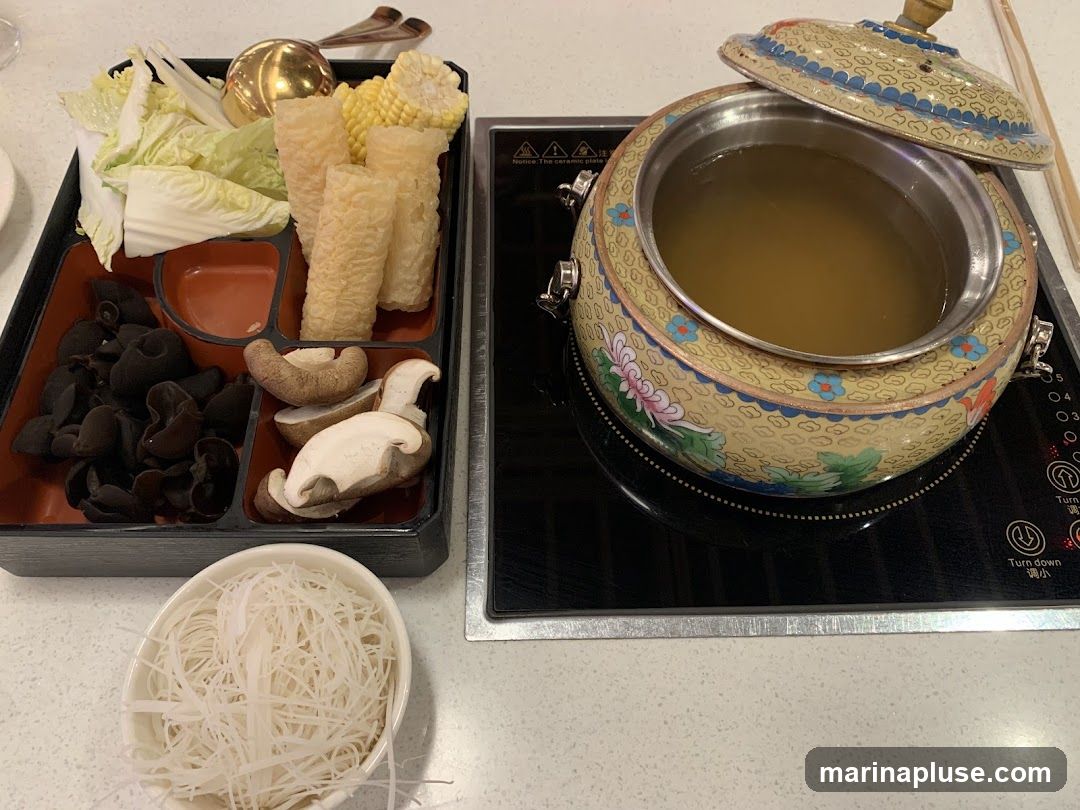 A hot pot setup with a decorative pot filled with broth, a tray of assorted ingredients including napa cabbage, corn, fried tofu puffs, wood ear mushrooms, and shiitake mushrooms, and a bowl of thin noodles. The pot is placed on an induction cooker.