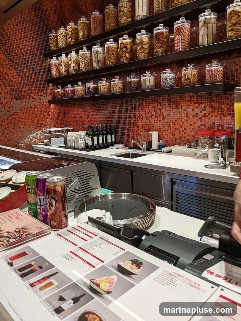 A counter displays a dessert menu with images of various desserts, including layered cakes and ice cream. To the left, there are cans of soda and a brochure titled 'Hotpot Bundle Deals'. Behind the counter, shelves are stocked with jars of cookies, crackers, and other snacks. A sink and bottles of wine are also visible.