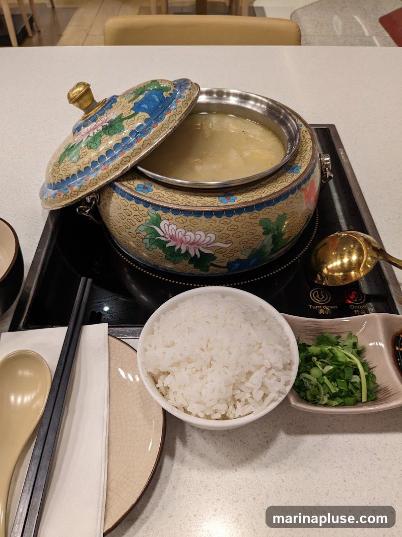 A decorative hot pot filled with broth sits on an induction cooker. Beside it is a bowl of white rice, a small dish of chopped green onions and cilantro, chopsticks, a spoon, and a napkin.