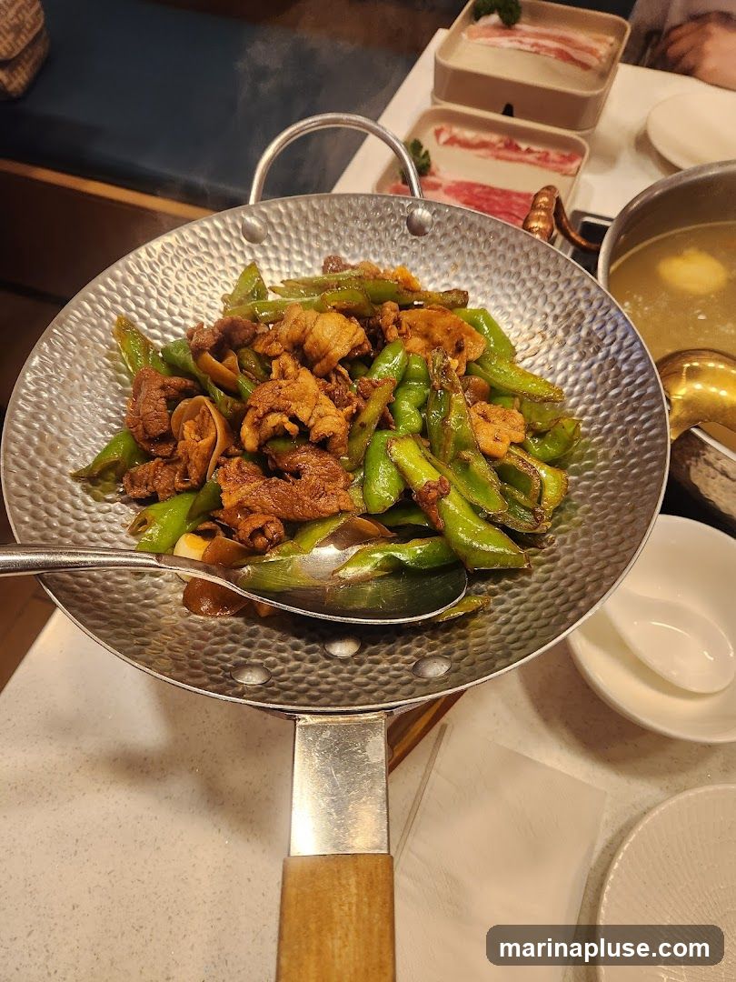 A close-up shot of a stir-fried dish served in a metal wok. The dish contains sliced meat, green peppers, and possibly other vegetables. Steam is rising from the wok, indicating it is hot. In the background, there are plates of raw meat, a pot of broth, and white bowls.