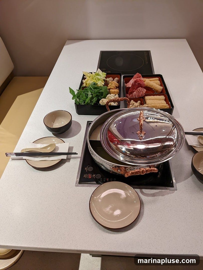 A table set for a hot pot meal with various ingredients including thinly sliced meat, leafy greens, and other vegetables. A pot with a lid sits on an induction cooker, ready to be heated. Plates, bowls, and chopsticks are arranged for diners.