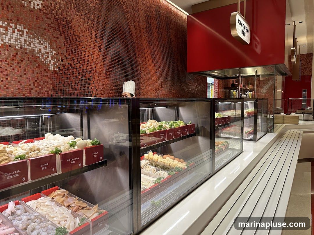 A display of various food items, including seafood, dumplings, and vegetables, arranged in refrigerated glass cases within a restaurant. A chef in a white hat is visible in the background. The restaurant features red tiled walls and red accents.