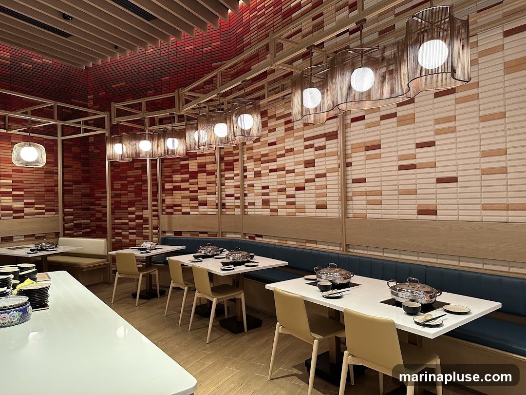 A modern restaurant dining area with tables set for hot pot. The walls are decorated with a mosaic of red, beige, and brown rectangular tiles. Pendant lights with woven shades hang from the ceiling. Booth seating lines the walls, with tables and chairs in the center.