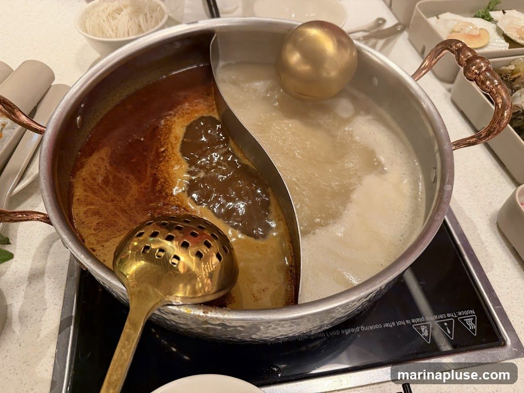 A split hot pot with two different broths, one spicy and one creamy, sits on an induction cooktop. A slotted spoon and a ladle are in the pot. In the background, a bowl of noodles and other dishes are visible.