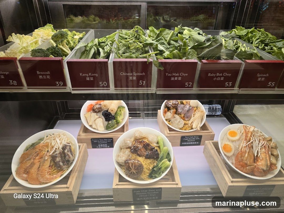 A display case filled with fresh vegetables and prepared noodle dishes. The vegetables are in individual containers with labels and prices. Below the vegetables are several bowls of noodles with various toppings, including shrimp, pork, and eggs.