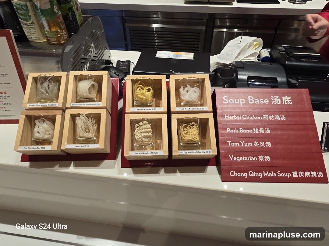 A display of various types of noodles in small glass containers, each labeled with its name in English and Chinese. Next to the noodles is a red sign listing different soup bases, also with English and Chinese names. Behind the display, a counter with a cash register and receipts is visible.
